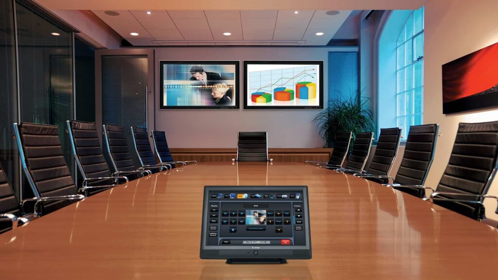 Boardroom-intergration-company Modern conference room with a long table, black leather chairs, dual wall-mounted screens displaying visuals, and a central touchscreen control panel.