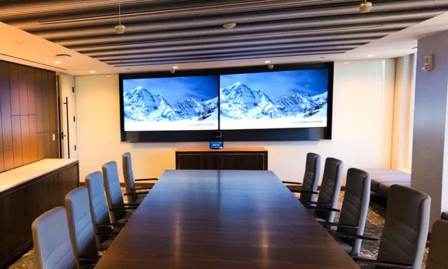 MicrosoftTeams-image-3-2-890x535 Contemporary conference room with a black table, several black chairs, and a large video wall displaying business visuals and city imagery, with a tablet and keyboard on the tabletop.