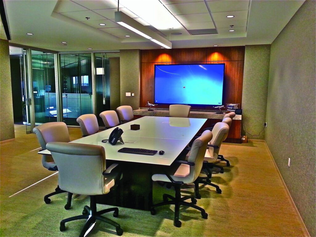 b2ap3_large_conference-room-av-considerations-for-the-modern-day Modern conference room with a large central table and multiple high-back chairs arranged neatly around it. The room features a minimalist design with clean lines, soft lighting, and a professional ambiance ideal for executive meetings, team collaborations, or video conferences.