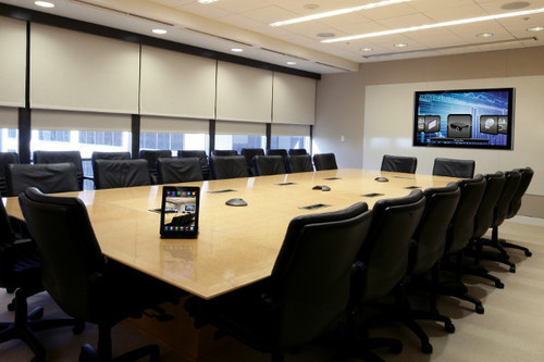conference-room1 Modern boardroom featuring a large central table, black mesh chairs, a wall-mounted multi-screen display showing charts, maps, and cityscapes, with a tablet and keyboard placed on the table.