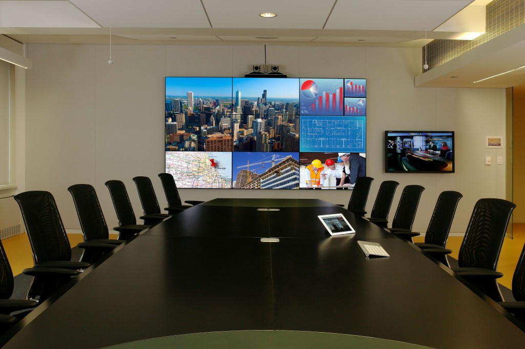 conference-room2 Sleek conference room with a long black table, ergonomic chairs, a large video wall displaying multiple visuals, and a tablet and keyboard on the table.