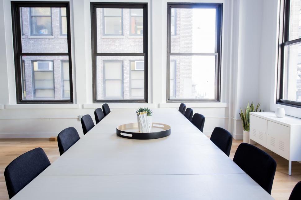 modern-office-conference-room-with-large-windows Spacious conference room featuring a long meeting table surrounded by comfortable office chairs. The room is well-lit with natural light from large windows and is equipped with modern decor, including wall art, plants, and professional lighting. Ideal for corporate meetings or team discussions in a clean, organized environment.